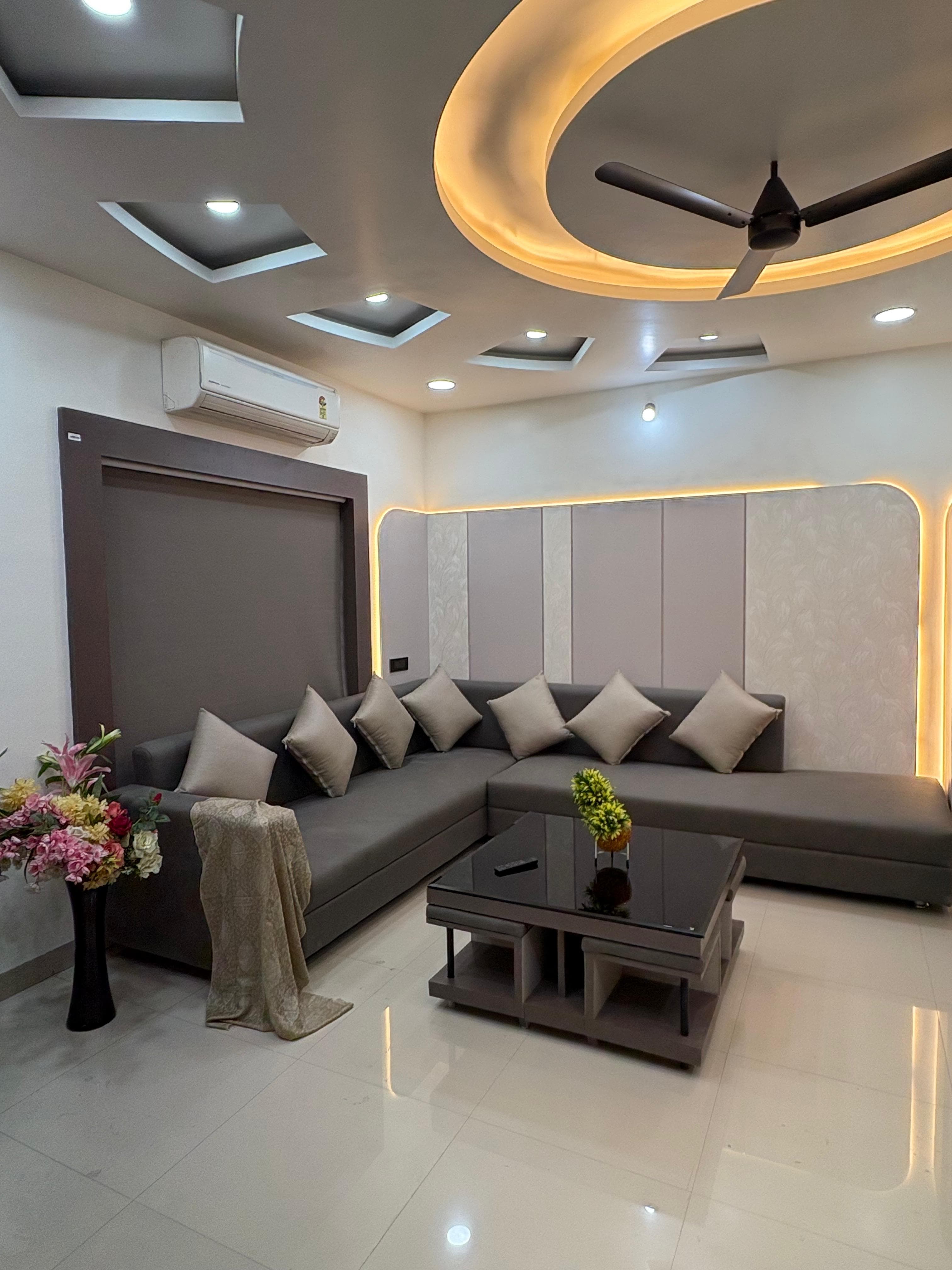 ग्रे सोफे, काली कॉफी टेबल और सुंदर फॉल्स सीलिंग लाइटिंग वाला आधुनिक लिविंग रूम।
